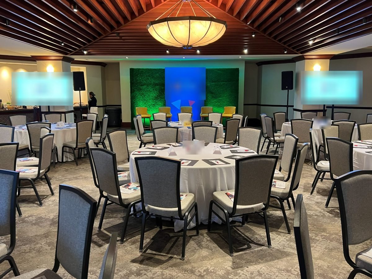Banquet room with round tables, two display screens on stands and a panel stage against a green-lit living wall