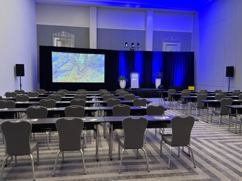 Classroom-style meeting room facing a projection screen, a podium and blue-lit black drape